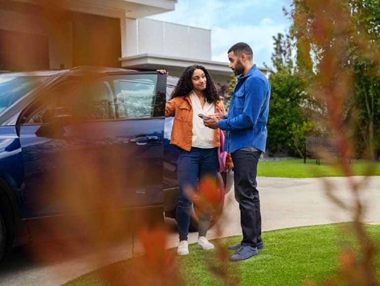 A couple stands by their car in a suburban driveway, discussing next to an open door.