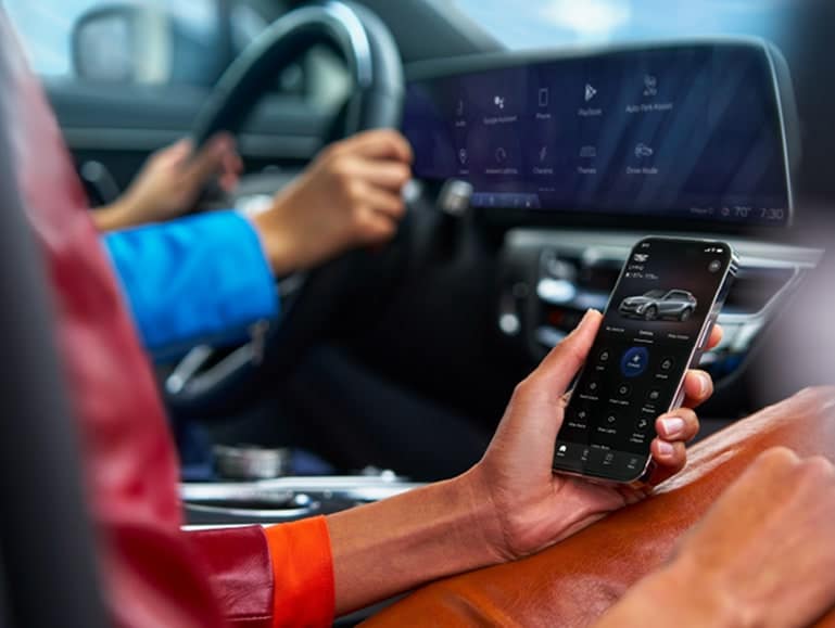 Person in car holds smartphone with vehicle control app open, dashboard visible.
