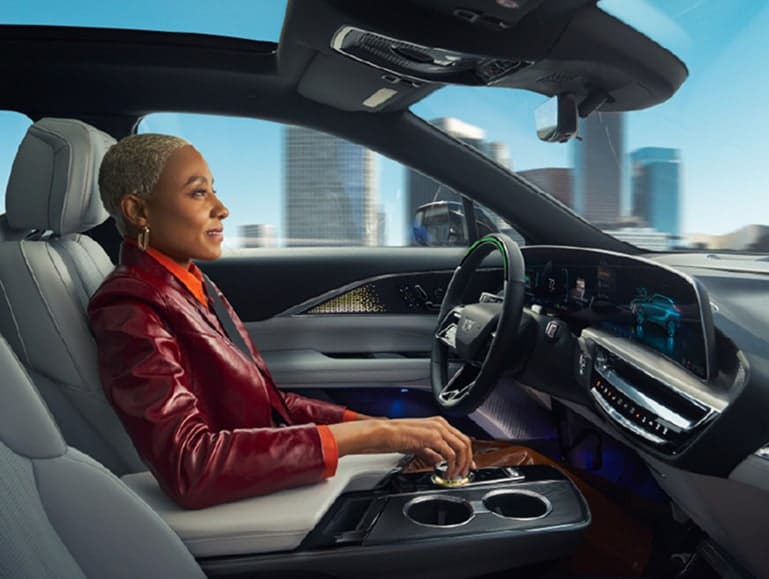 Woman driving a modern car with advanced dashboard display, cityscape visible through windows.