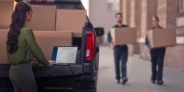 Woman using a laptop on a truck tailgate while two people carry boxes in an alleyway.