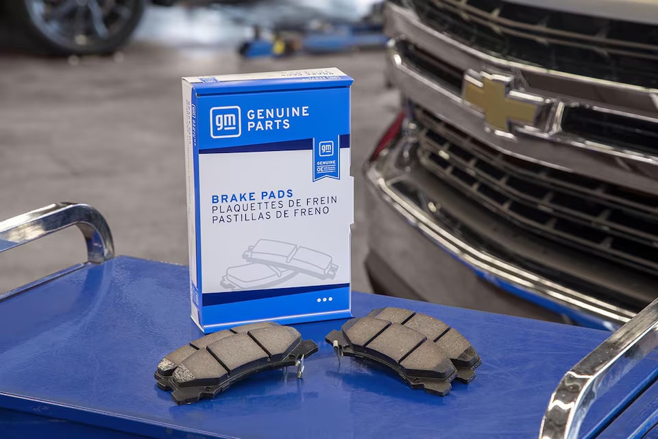 GM Genuine Parts brake pads on a blue cart near a Chevrolet vehicle grille.