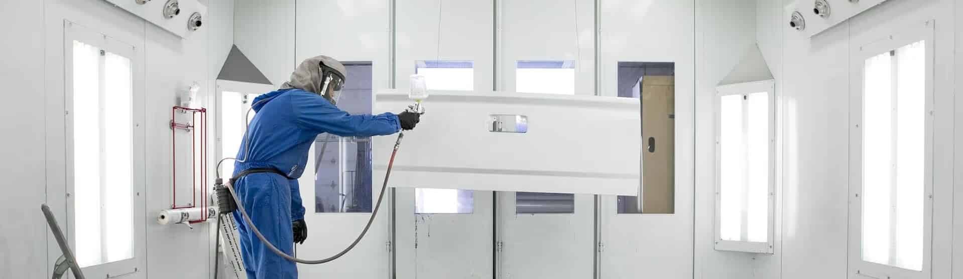 Worker in protective gear spray-paints a vehicle door in a well-lit industrial booth.