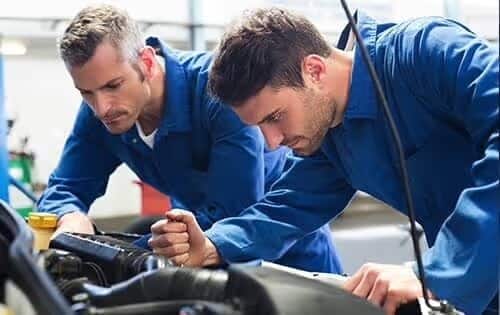 Two mechanics in blue coveralls work intently on a car engine in a garage setting.
