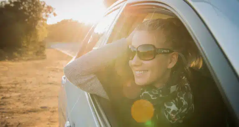 Woman enjoying a sunny road trip, smiling while leaning out car window, wearing sunglasses.