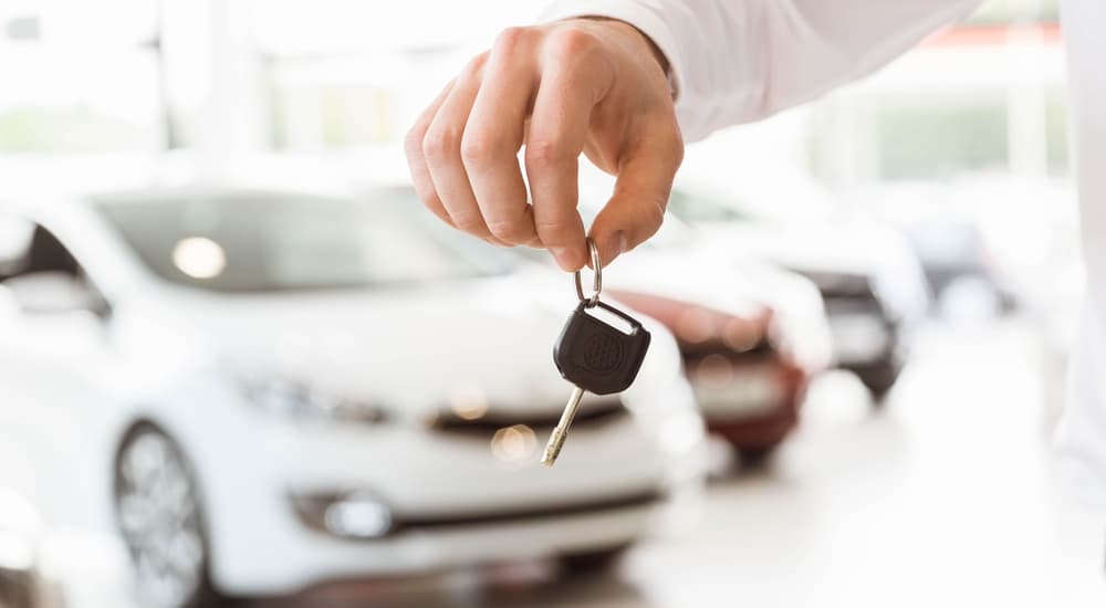 Hand holding car key in dealership with blurred cars in the background.