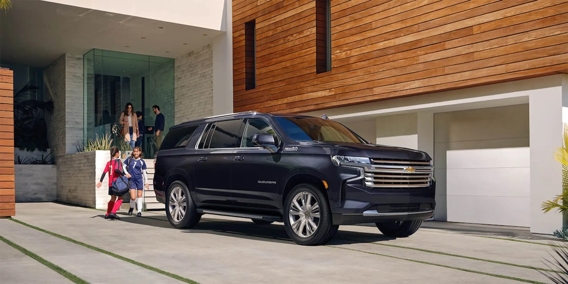 A family heading out from their home beside a dark Chevrolet Suburban