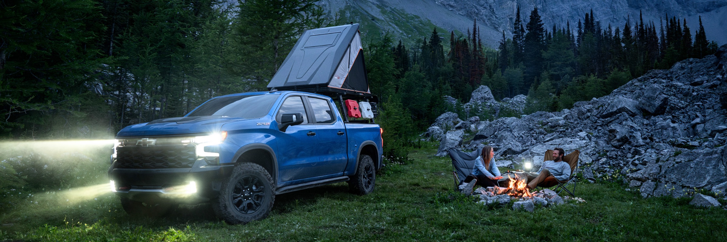 A blue Chevrolet Silverado ZR2 with a rooftop tent parked at a forest campsite at dusk, with two people sitting by the campfire