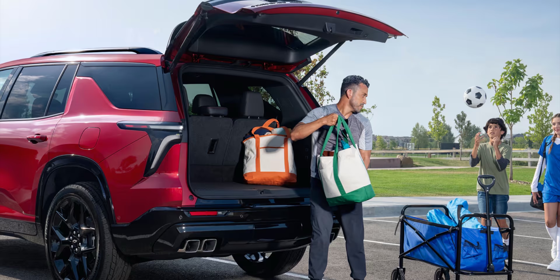 A father loading sports bags into the cargo area of a red Chevrolet Traverse while his kids play soccer